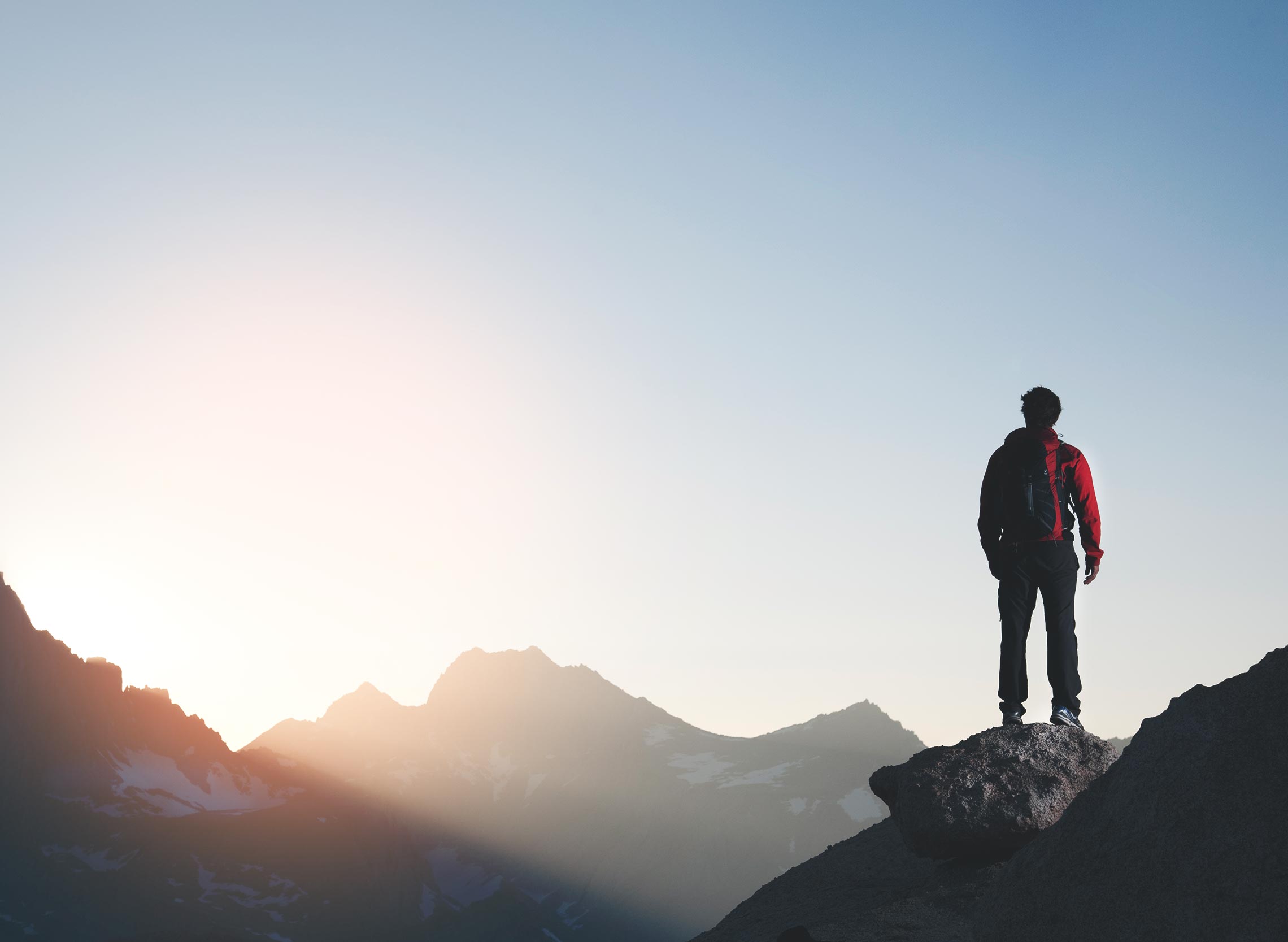 Silhouette of a person in a red and dark jacket standing on a rocky ridge overlooking mountains at sunrise, warm and cool tones