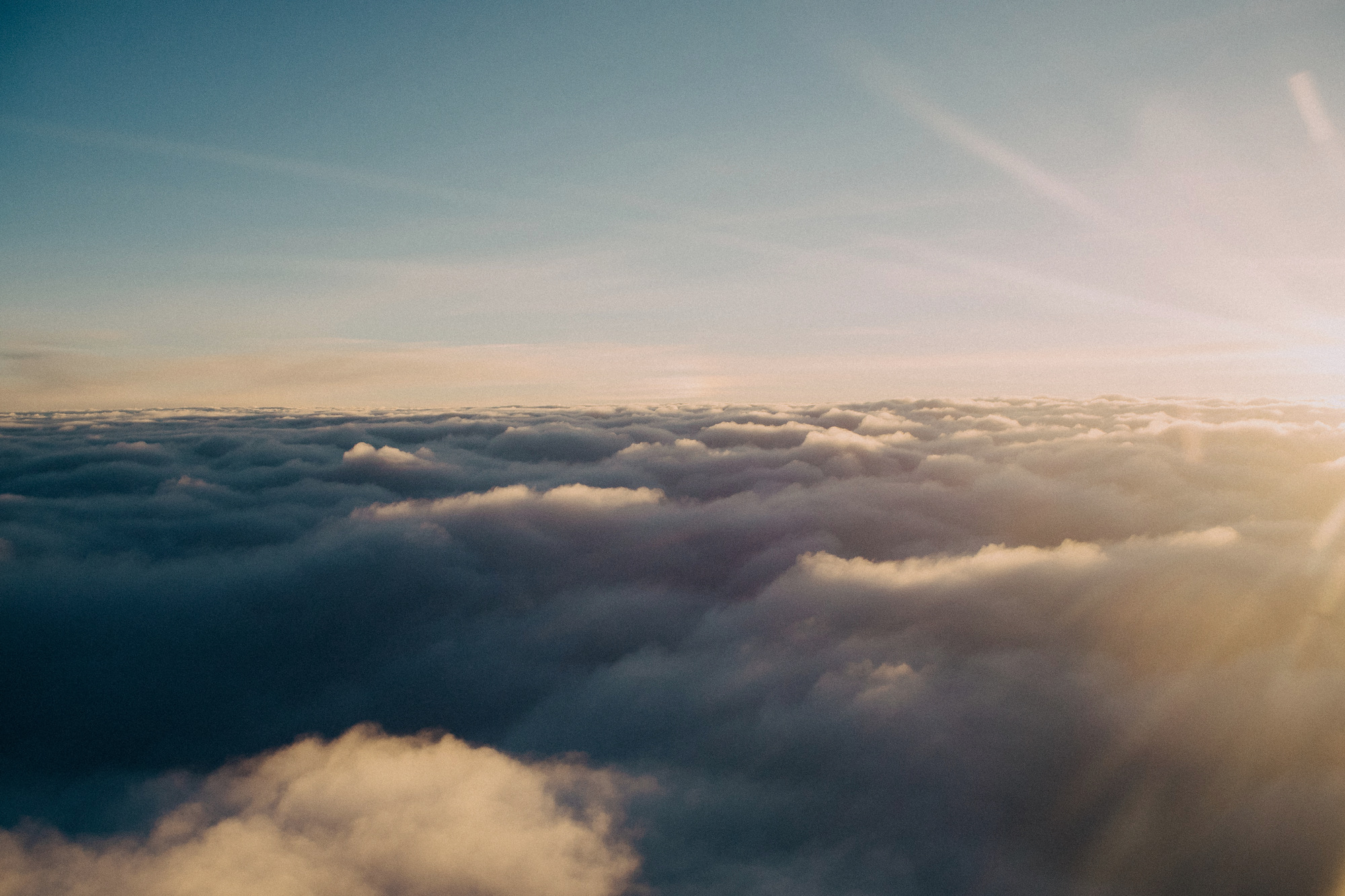 Aerial view above a thick layer of clouds lit by golden sunlight, blue and orange tones, serene expansive mood